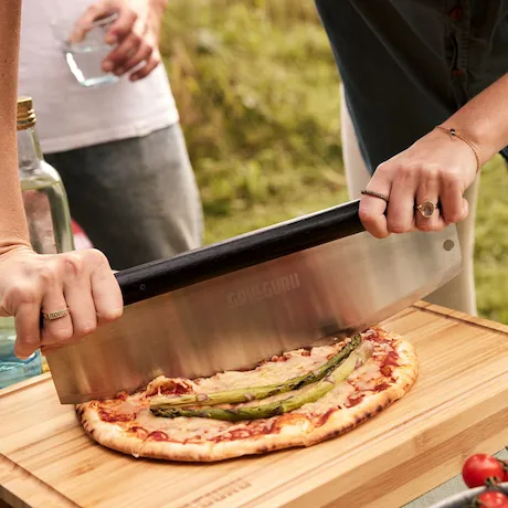 Eine Person schneidet eine Pizza mit Spargel auf einem Holzbrett mit einem großen Pizzaschneider in einer Outdoor-Umgebung.